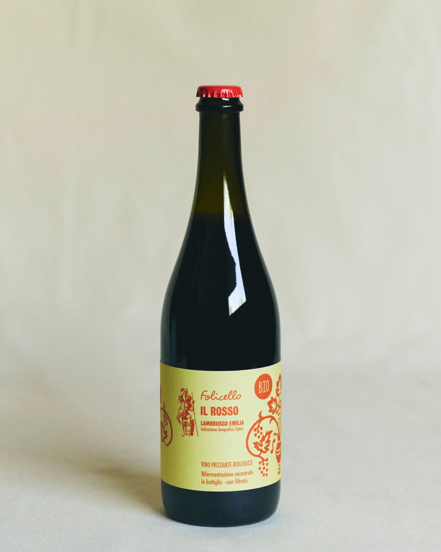 A transparent bottle containing red wine, sealed with a metal crown cap. The yellow label reads “Folicello Il Rosso” and features illustrations of grapes and flowers in red lettering.
