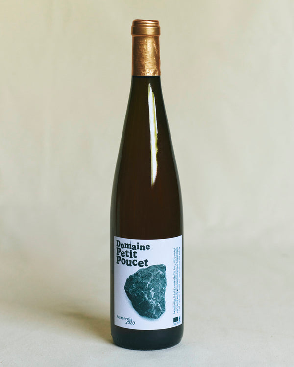 A slender brown Alsace wine bottle with a minimalist white label bearing the words “Petit Poucet” alongside a delicate white illustration featuring a stone and the text “Auxerrois 2020.”
