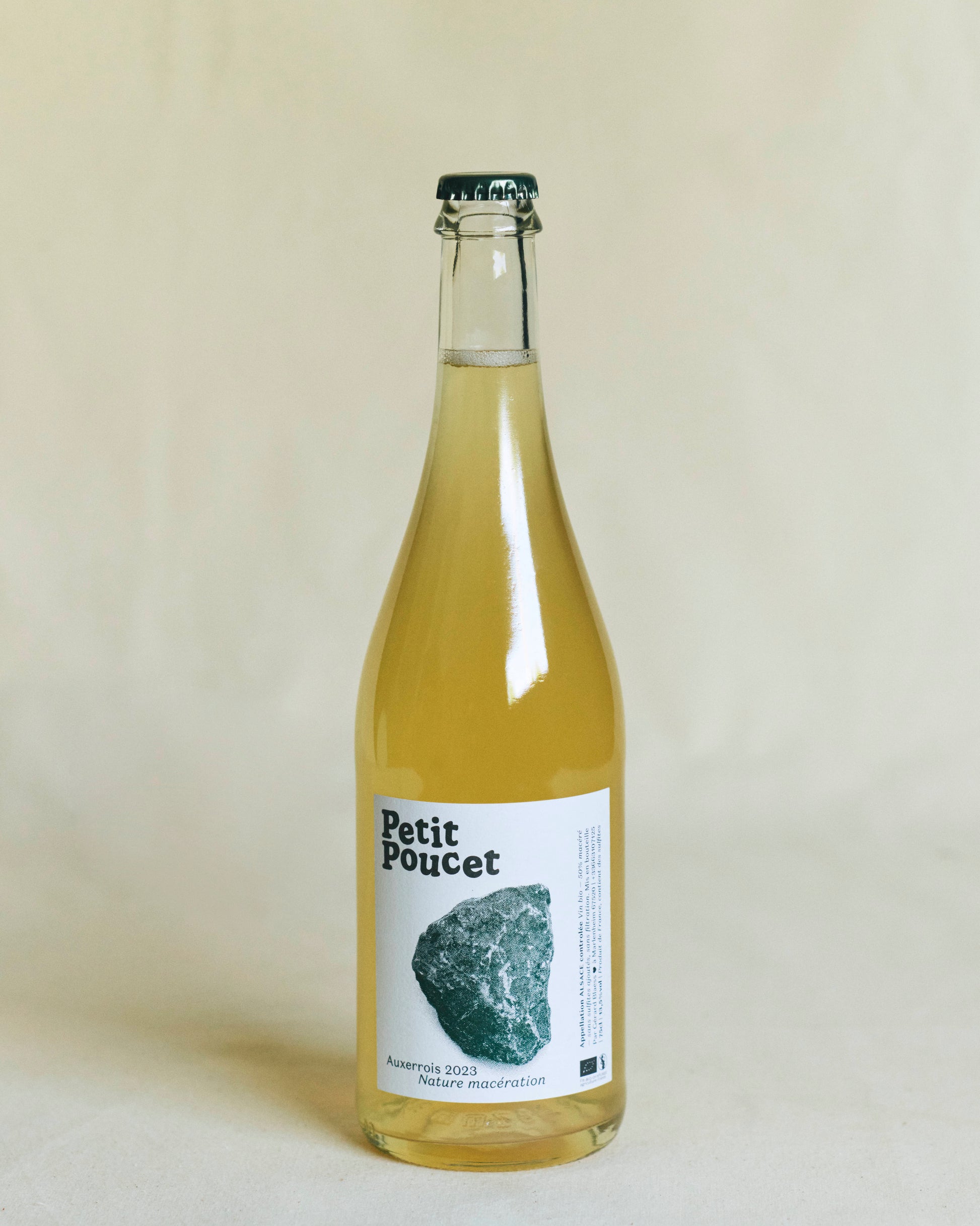 A beautiful transparent white wine bottle with a minimalist white label bearing the words “Petit Poucet” next to a soft white illustration featuring a stone and the text “Auxerrois 2023 Nature Maceration.”