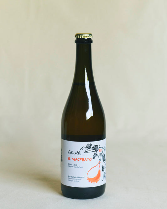 A brown bottle of wine, sealed with a metal crown cap. The white label reads “Folicello Il Macerato” and features illustrations of grapes and flowers in orange lettering.