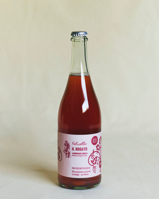 A transparent bottle containing rosé wine, sealed with a metal crown cap. The pink label reads “Folicello Il Rosato” and features illustrations of grapes and flowers.