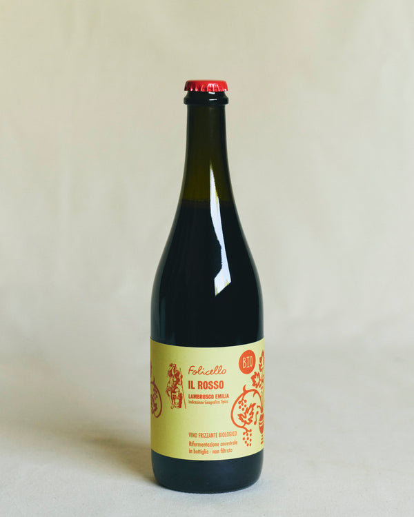 A transparent bottle containing red wine, sealed with a metal crown cap. The yellow label reads “Folicello Il Rosso” and features illustrations of grapes and flowers in red lettering.